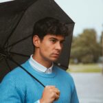 A thoughtful man in blue sweater holding an umbrella on a rainy Munich day.